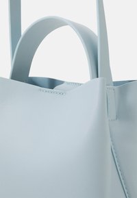 Primo piano di una borsa in pelle blu chiaro che mostra una superficie liscia, dettagli di cucitura e due manici su uno sfondo chiaro.
