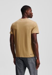 Korte mouwen, beige t-shirt van zachte katoen. Bevat een ronde halslijn en een klein logopaneel aan de zoom. Gepaard met grijze jeans.