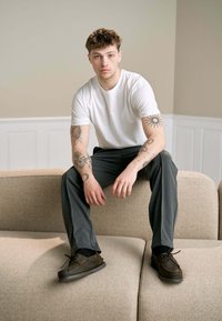 Hvid crew-neck t-shirt, grå bukser og brune snørebåndssko. Sidder på en beige polstret sofa med en ru overflade, neutrale farver.