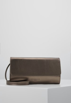 Petit sac bandoulière en cuir marron avec fermeture à rabat et bandoulière fine, posé sur une surface blanche devant un fond uni.
