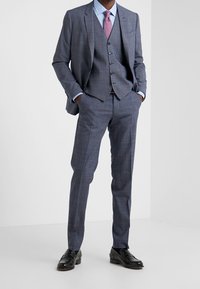 Man wearing a blue checked three-piece suit with a lavender tie and black dress shoes, standing with hands in pockets.