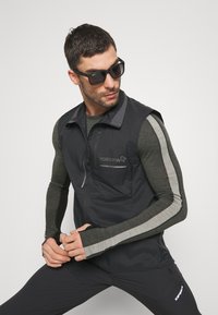 Czarna kamizelka z zamkiem błyskawicznym z szarymi akcentami i logo. Model ma na sobie szarą koszulę z długim rękawem i czarne spodnie oraz okulary przeciwsłoneczne.