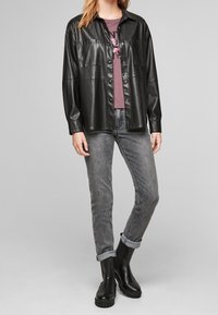 Černá kožená košile s kapsami na hrudi, nošená přes vínově červené tričko s grafikou. V kombinaci se šedými džínami a černými kotníkovými botami.