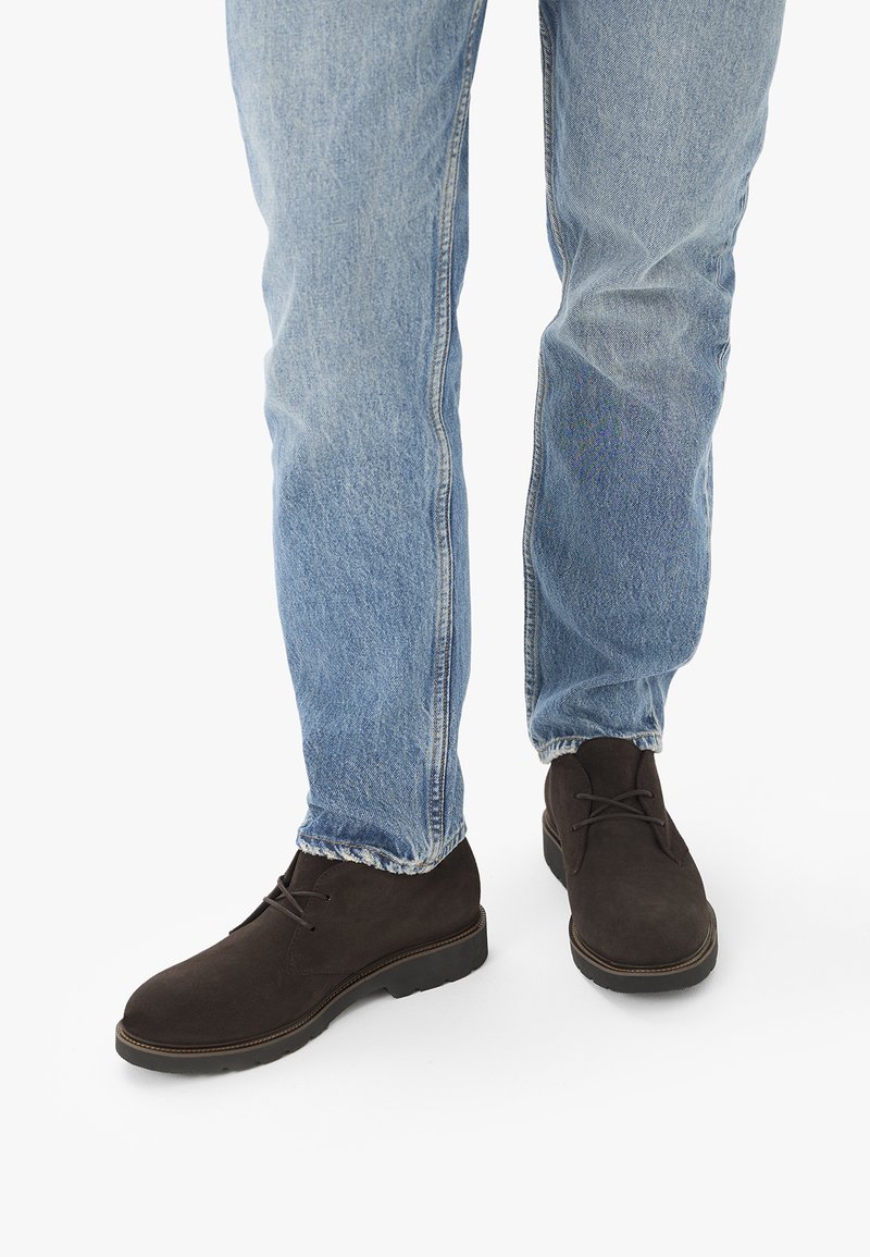 Personne portant un jean bleu clair et des bottines en daim marron foncé à lacets, debout sur un fond blanc.