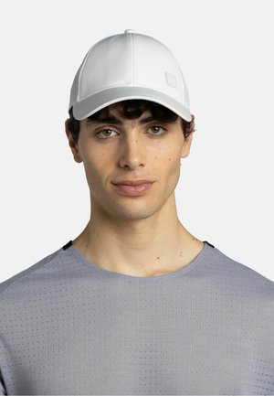 Joven con ojos marrones que lleva una gorra blanca y una camiseta deportiva perforada gris claro, mirando al frente sobre un fondo liso.