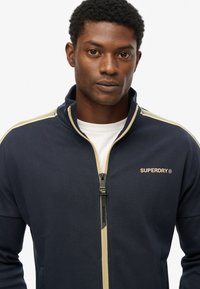 Veste zippée bleu marine en mélange de coton avec un col montant. Présente des rayures beiges sur les épaules et un logo sur la poitrine.