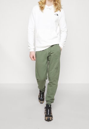 Bílá mikina s kapucí a přední kapsou s logem, spárovaná se zelenými tepláky s logem a černými sportovními botami se žlutými akcenty.