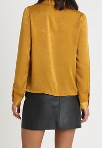 Blouse en soie jaune moutarde à manches longues, à coupe droite et avec des poignets à boutons, associée à une jupe en simili cuir noire.