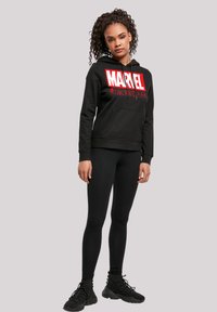 Black hoodie with a prominent red "MARVEL" logo, kangaroo pocket, and ribbed cuffs, paired with black leggings and athletic shoes.