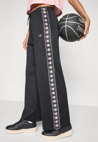 A pessoa veste calças de treino pretas da Champion e ténis pretos, segurando uma bola de basquetebol preta contra um fundo simples.
