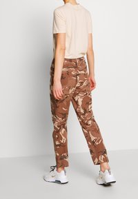 Bruine camouflagebroek met een tapered fit, zakken en knoopaccenten, gecombineerd met een beige T-shirt en witte sneakers.