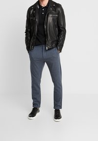 Mies, joka käyttää mustaa nahkatakkia, mustaa poolopaitaa, sinisiä housuja ja mustia lenkkareita, joissa on valkoiset pohjat, seisoo kädet taskuissa.