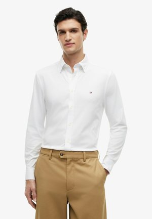 Homme debout, une main dans la poche, portant une chemise blanche à boutons et un pantalon beige sur un fond blanc uni.