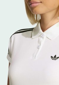 Biela polo košeľa s čiernym trojprúžkovým dizajnom na pleciach, rebrovaný golier, dva gombíky na zapínanie a malý čierny logotyp na hrudi.