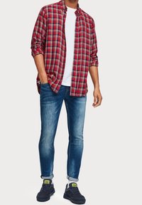 Rood en marineblauw geruite flanellen shirt, witte t-shirt eronder, blauwe slim-fit jeans en donkere sneakers met grijze en groene accenten.