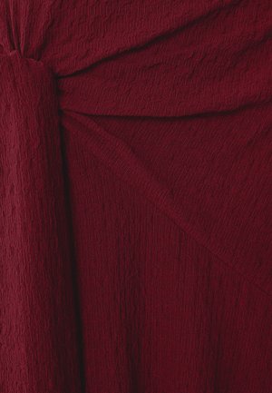 Tessuto bordeaux texturizzato con una superficie arricciata, caratterizzato da un dettaglio attorcigliato su un lato e da una caduta morbida, offrendo profondità visiva.