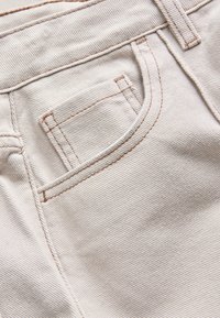 Close-up of light beige denim fabric showing the front pocket and visible brown stitching details.