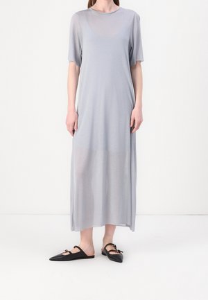 Kobieta w jasnoszarej, krótkorękawowej sukience sięgającej kostek, wykonanej z przezroczystej tkaniny, w połączeniu z czarnymi płaskimi butami na szpic z paskami.