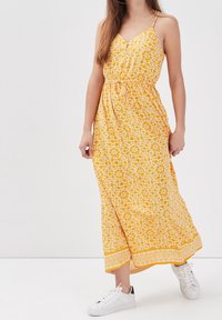 Robe maxi jaune à motif floral, bretelles spaghetti réglables, taille élastique et ourlet à motif contrasté. Portée avec des sneakers blanches.
