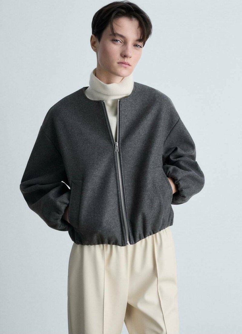 Giacca bomber grigia con zip e vestibilità comoda, caratterizzata da un collo alto e orlo elasticizzato. Indossata sopra un dolcevita crema. Tessuto testurizzato.