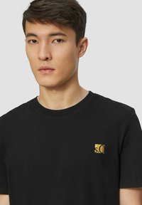 Young man with short dark hair wearing a black t-shirt with a small gold "s.Oliver" logo on the chest, neutral expression, light background.