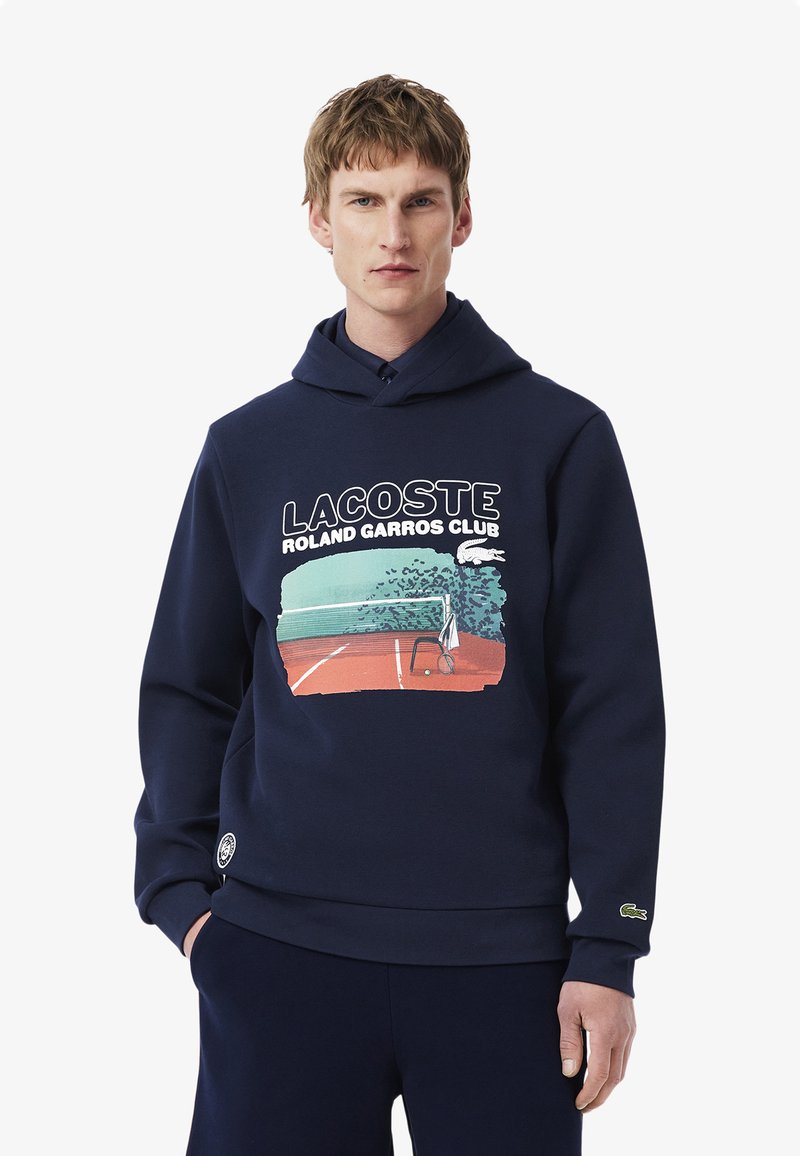 Marinblå sweatshirt med en framsidesgrafik av en tennisscen, med en dragslundshuva och en liten grön logotypaccent på fållen. Mjuk textur.