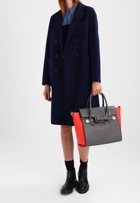 Szara i czerwona torebka o strukturalnym designie, z akcentem w postaci kokardy, podwójnymi uchwytami oraz gładką fakturą. Modelka nosi granatowy płaszcz.