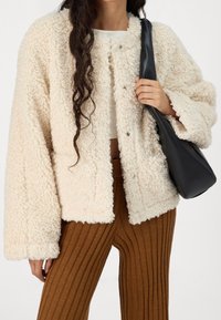 Beige, fluffig, kort jacka med en texturerad finish, med tryckknappar. Kombinerad med ribbade bruna byxor och en svart axelväska.