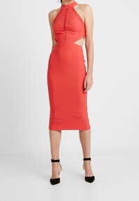Červené bezramínkové šaty s halter krkem, zdobené krajkovými detaily, s výřezy na bocích a přiléhající midi délkou. Hladká textura a bodycon silueta.
