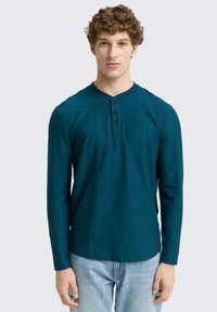 Türkises Langarm-Henley-Shirt mit rundem Ausschnitt, drei schwarzen Knöpfen und strukturiertem Stoff, kombiniert mit hellblauen Jeans.