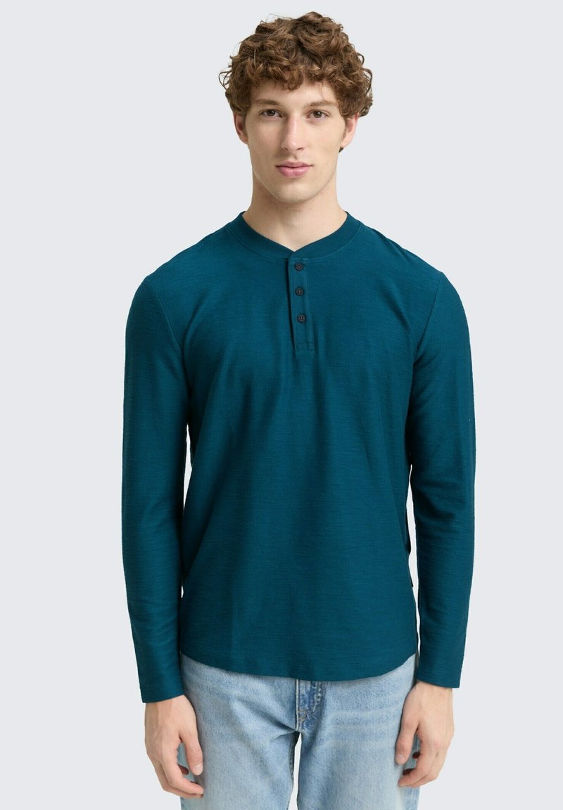 Türkises Langarm-Henley-Shirt mit rundem Ausschnitt, drei schwarzen Knöpfen und strukturiertem Stoff, kombiniert mit hellblauen Jeans.