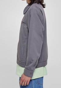 Šedá lehká bunda s límcem, kapsou na zip a manžetami na knoflíky, nošená přes světle zelenou košili a modré džíny. Jednoduchý a funkční design.