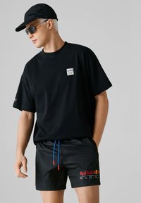 Moški z črno kapico Red Bull Racing, sončnimi očali, preveliko majico in kratkimi hlačami z modrimi vrvicami na enobarvnem sivem ozadju.