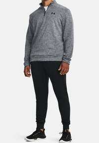 Grå quarter-zip pullover med en tekstureret overflade og sort logo, parret med sorte tapered bukser og sorte træningssko med hvide såler.