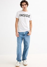 Camiseta blanca de manga corta con texto negro que dice "INSIDE", combinada con unos vaqueros holgados de un azul claro y zapatillas grises con detalles blancos.
