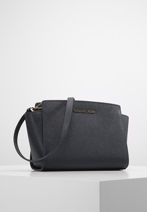 Czarna torebka na ramię z fakturowanej skóry z złotymi elementami i regulowanym paskiem na białym postumencie.