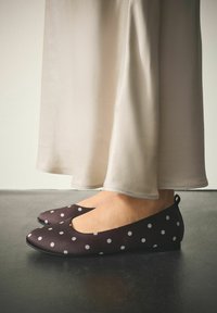 Brązowe baleriny w groszki z zaokrąglonym noskiem i niskim profilem, zestawione z lekką, jasnobeżową tkaniną. Widoczna teksturowana powierzchnia.