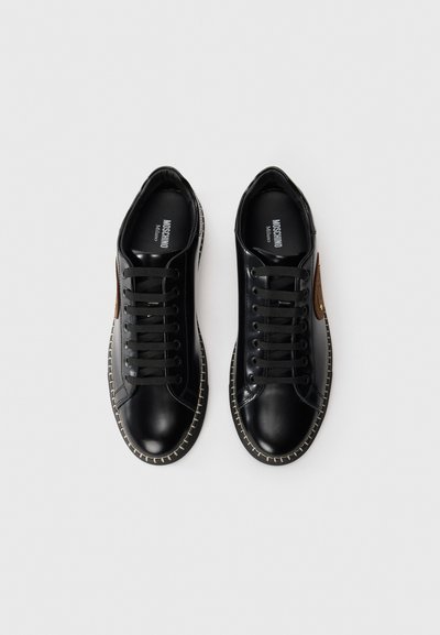 Schwarze Leder-Sneaker mit runder Spitze, schwarzen Schnürsenkeln und einem kontrastierenden beigen Stich entlang der Sohle. Mit einem braunen Logoaufnäher an der Seite.