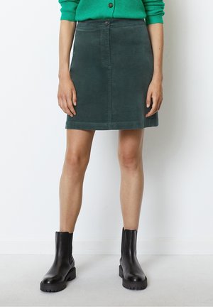 Kvinde iført en mørkegrøn fløjlsmininederdel, grøn knap-sweater og sorte chunky ankelstøvler stående mod en hvid baggrund.