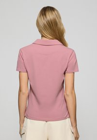 Pikétröja i pastellrosa, med korta ärmar, krage och ett mjukt, stretchigt tyg. Bakifrån visas en enkel, ren design.