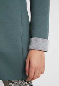 Close-up van een hand die naast zich rust, terwijl deze een groen langemouwen shirt met een grijze manchet en zwart-witte gestreepte broek draagt.