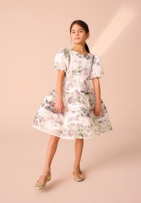 Cvetlična obleka z napihnjenimi rokavi, večplastno krilo in mehak material. Barve vključujejo belo, zeleno in vijolično. Na pasu ima cvetlični okrasek.