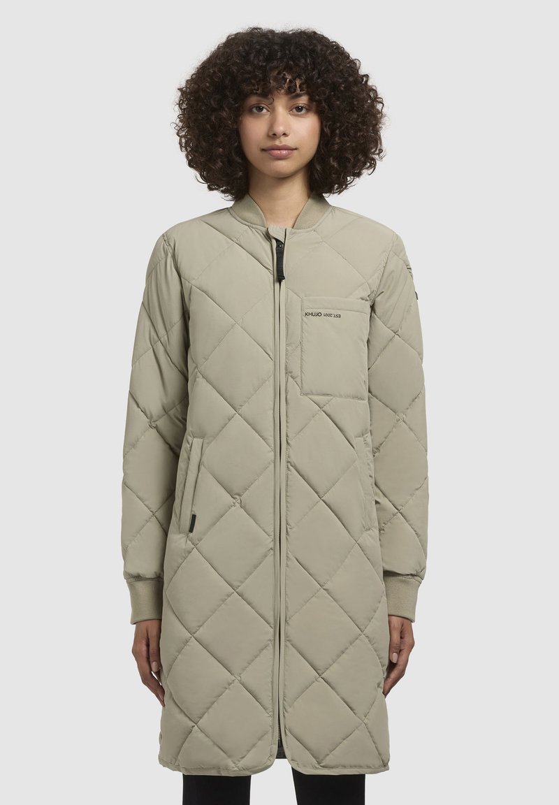 Cappotto lungo trapuntato di colore beige chiaro con polsini a costine, chiusura con zip, tasche laterali e una tasca sul petto con dettagli del logo.