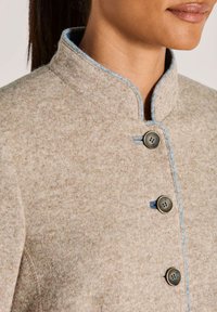 Close-up van een beige wollen jas met blauwe accenten en drie donkere knopen, gedragen door een persoon met donker haar dat teruggebonden is.