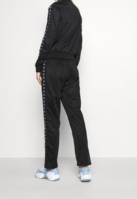 Svart träningsdräkt med en zip-up jacka och elastiska byxor, med logomönster längs ärmarna och sidorna. Kombinerad med blå sneakers.