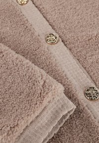 Beige fleece vest met een gestructureerde stof, voorzien van een gladde gebreide rand en gouden decoratieve knopen. Zacht, warm en gezellig ontwerp.