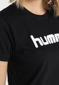 Camiseta de algodón negra con un logo blanco de "humm". El cuello redondo y las mangas cortas crean un diseño simple y casual. Textura suave.