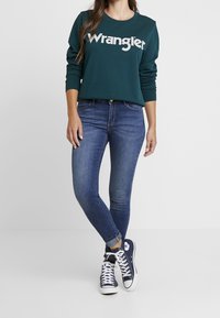 Sudadera verde oscuro con el logotipo de "Wrangler", jeans ajustados en azul y zapatillas de deporte en color marino. El conjunto presenta un look casual con los puños arremangados.