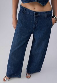 Laiade lõikega tumesinised teksapüksid, millel on vertikaalsed hõbedased helmestikest kaunistused, kantud hõbedaste avatud varbaga kontsakingade ja hõbedase käevõruga.