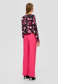 Mujer de espaldas con zapatos negros de tacón, pantalones anchos de pierna ancha en color rosa brillante y una blusa negra con estampados florales en rosa y blanco.